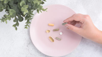 Pink plate with assortment of pill supplements on top with manicured hand picking up one of the pills with eucalyptus plant nearby on table with plate